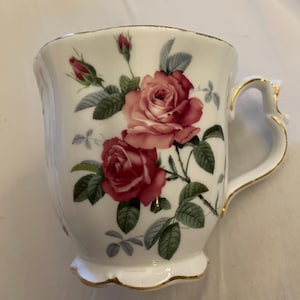 May include: A white porcelain mug featuring a floral pattern of pink and red roses with green leaves and buds. The mug has a scalloped base and rim, both accented with a thin gold trim, and a curved handle with gold detailing along the edges.