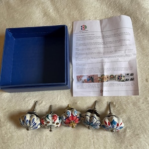 May include: Five ceramic drawer knobs with multi-colored floral patterns, a blue storage box, and an instruction sheet titled How to Install a Drawer Knob by IBP by Ibacrafts. Each knob features a metal bolt and a rounded, hand-painted design.