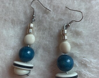 Può includere: Un paio di orecchini pendenti con ganci color argento, piccole perline distanziatrici metalliche, perline rotonde color crema, perle sferiche più grandi color blu ottanio e dischi inferiori con strisce orizzontali color crema e nero.
