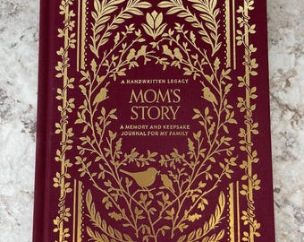 Può includere: Un diario con copertina rigida color bordeaux intenso con dettagli in lamina d'oro. La copertina presenta intricate illustrazioni botaniche di rami e uccelli, con il testo A HANDWRITTEN LEGACY MOM'S STORY A MEMORY AND KEEPSAKE JOURNAL FOR MY FAMILY stampato in oro.