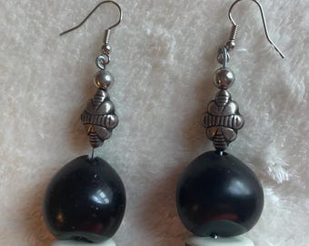 Può includere: Un paio di orecchini pendenti con ganci in metallo argentato e una perlina decorativa a forma di ape. Sotto l'ape, ogni orecchino presenta una grande perlina nera liscia, un disco bianco piatto con una striscia nera centrale e una piccola perlina grigio-bianca.