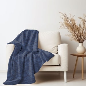 May include: A blue throw blanket with a wavy pattern draped over a white armchair. The blanket is made of soft, plush fabric and has a contemporary design.