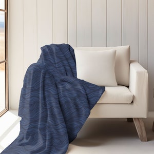 May include: A blue throw blanket with a wavy pattern draped over a white couch. The couch has a white pillow and is in a room with a window and a wooden side table.
