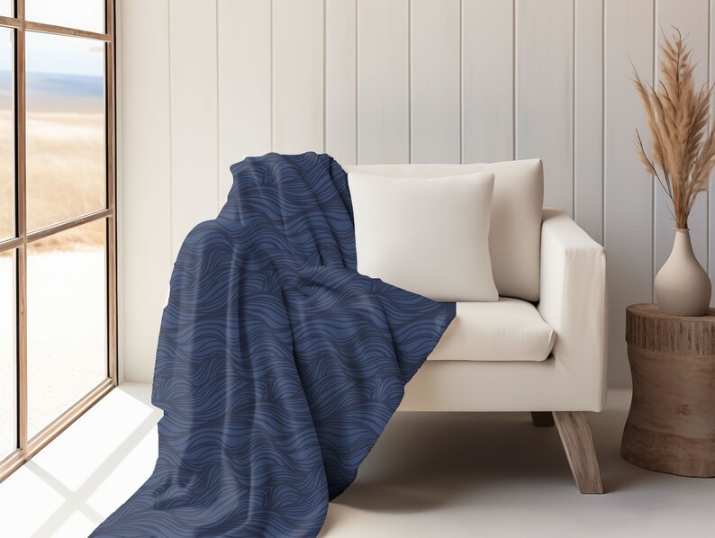 May include: A blue throw blanket with a wavy pattern draped over a white couch. The couch has a white pillow and is in a room with a window and a wooden side table.