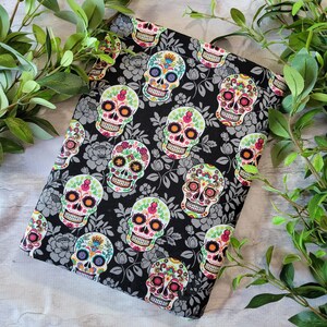 May include: Black fabric with a repeating pattern of colorful sugar skulls on a black background. The skulls have floral details and are surrounded by gray floral designs.