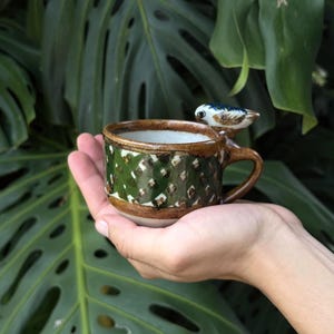 May include: A small ceramic cup featuring a dark green exterior with a textured diamond pattern and a brown rim. A sculpted, hand-painted bird figure with white, blue, and brown details is attached to the top edge of the cup near the handle.
