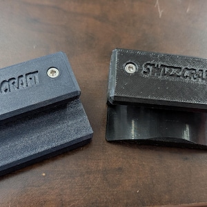 May include: Two black and dark gray rectangular objects with the text "SWIZZCRAFT" are displayed on a wooden surface. Each object has two silver screws. One object is stacked on top of another.