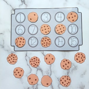 May include: A cookie-themed counting activity with a baking sheet with 15 numbered circles and 15 individual cookies. The numbers 1, 3, 4, 6, 7, 10, 12, 14, and 15 are visible on the baking sheet.
