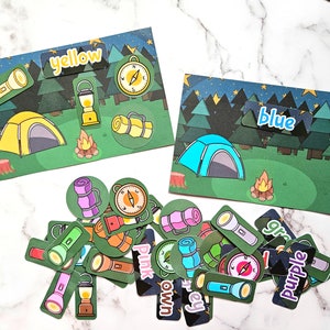 May include: A camping-themed educational game with colourful illustrations. Features cards with tents, campfires, and flashlights, labelled with colours like yellow and blue. Includes matching game pieces.
