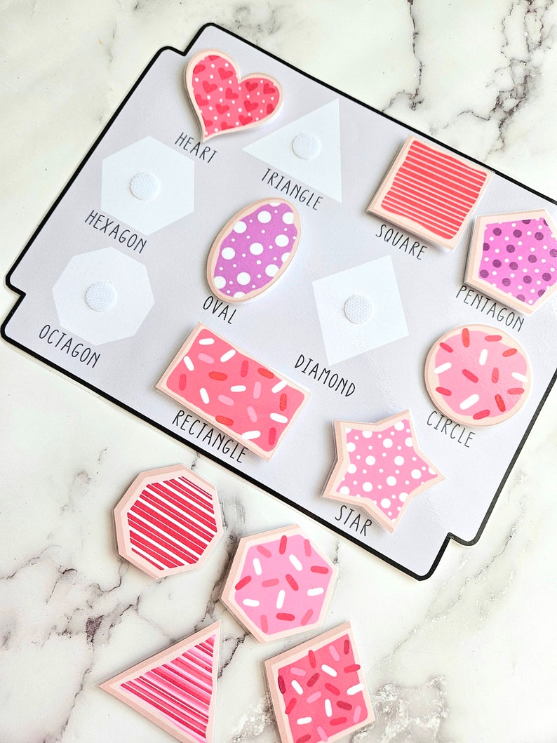 Pode incluir: Um conjunto de 10 formas de feltro coloridas com padr&otilde;es rosa e branco. As formas incluem um cora&ccedil;&atilde;o, um tri&acirc;ngulo, um quadrado, um oval, um pent&aacute;gono, um hex&aacute;gono, um oct&oacute;gono, um diamante, um c&iacute;rculo e uma estrela. Cada forma tem uma forma de feltro branca correspondente com um ponto de velcro.
