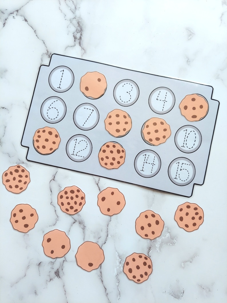 May include: A cookie-themed counting activity with a baking sheet and 15 cookies. The baking sheet has numbers 1 through 15 printed on it. The cookies are brown with chocolate chips and are scattered around the baking sheet.