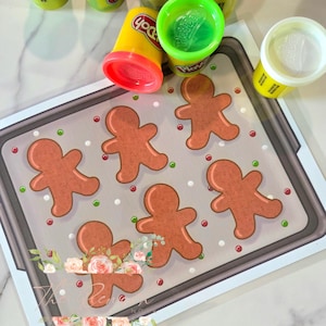 May include: A Play-Doh set with various colors, including green, blue, purple, and black, alongside a tray with gingerbread men. The tray has a festive design with red and green accents, suggesting a holiday theme.