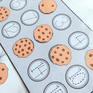 May include: A gray and white learning activity with a cookie theme. The activity has 12 circles with dotted lines for tracing numbers. There are 10 brown cookies with chocolate chips on top.