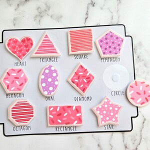 Pode incluir: Um quadro branco com formas rosa e vermelhas com nomes por baixo. As formas incluem um cora&ccedil;&atilde;o, um tri&acirc;ngulo, um quadrado, um pent&aacute;gono, um hex&aacute;gono, um oval, um diamante, um c&iacute;rculo, um oct&oacute;gono, um ret&acirc;ngulo e uma estrela.