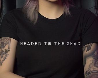 May include: A woman with purple hair wearing black headphones and a black t-shirt with the text "Headed to the Shad" sits in a black gaming chair. She has a tattoo on her left arm.