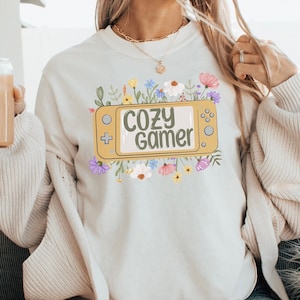 May include: A white sweatshirt featuring a graphic of a yellow video game controller with the text "Cozy Gamer" in green letters. The graphic is surrounded by colourful flowers.