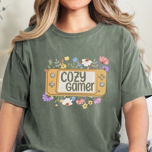 May include: A green t-shirt featuring a floral design and the text "Cozy Gamer" within a yellow video game controller graphic.