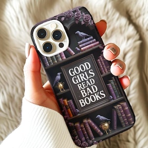 May include: A phone case featuring a bookshelf design with the text "GOOD GIRLS READ BAD BOOKS." The case has a dark background with purple roses, books, and decorative elements. The case is held by a person.