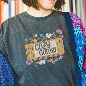 May include: A dark grey t-shirt featuring a floral design and a stylised video game controller with the text "Cozy Gamer" on it.