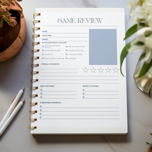 May include: A white notebook with a gold spiral binding. The cover has the text "GAME REVIEW" in black and gray. The notebook has sections for game details, overall thoughts, best features, worst features, and streamable moments. There are five stars for rating the game.