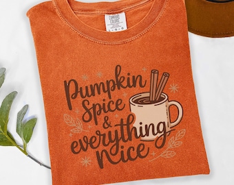 Może przedstawiać: Pomarańczowa koszulka Comfort Colors z napisem "Pumpkin Spice & Everything Nice" w ozdobnej czcionce. Projekt zawiera filiżankę kawy z laskami cynamonu. Brązowy kapelusz jest widoczny w tle.