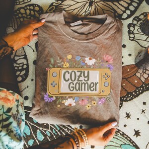 May include: A brown t-shirt featuring a yellow video game controller graphic with the text "Cozy Gamer" surrounded by flowers.