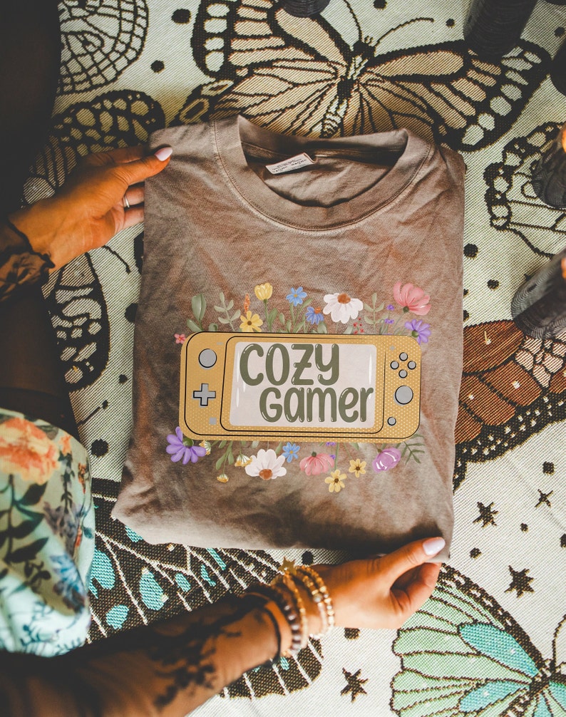 May include: A brown t-shirt featuring a yellow video game controller graphic with the text "Cozy Gamer" surrounded by flowers.
