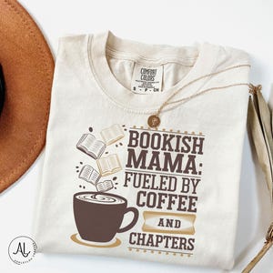 May include: Cream-colored t-shirt with the text "Bookish Mama: Fueled by Coffee and Chapters" in brown. The design includes a coffee cup and floating books. A gold necklace and a brown hat are also visible.