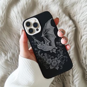May include: Black phone case featuring a detailed illustration of a bat with outstretched wings, surrounded by floral elements. The design is in shades of gray against a black background. The phone case has a white camera module.