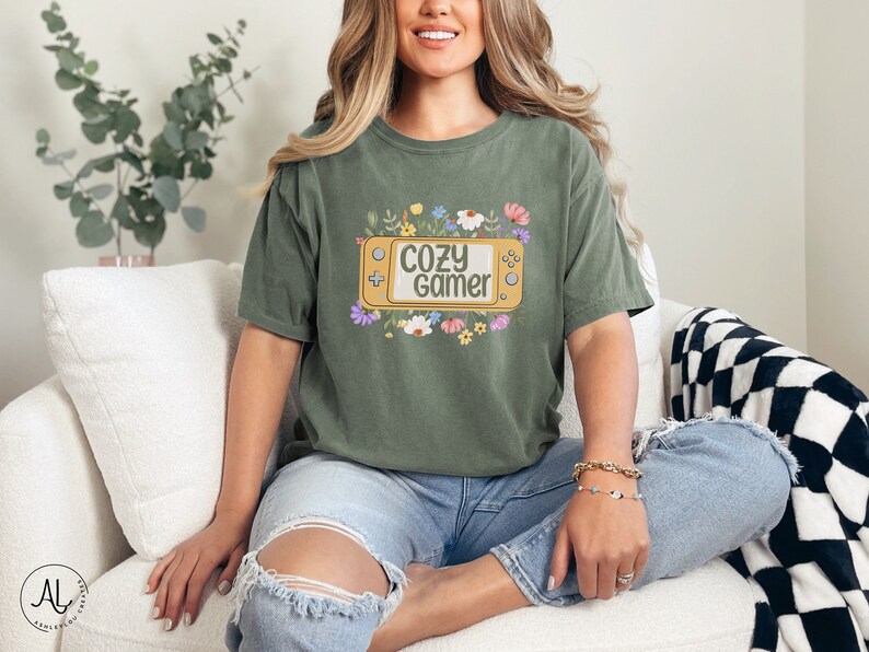 May include: A green t-shirt featuring a floral design and the text "Cozy Gamer" within a yellow video game controller graphic.