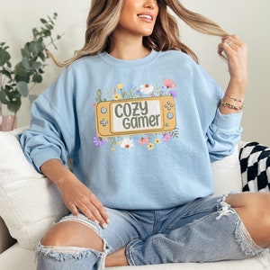 May include: A light blue sweatshirt with a graphic of a yellow video game console with the text "Cozy Gamer" surrounded by colorful flowers.