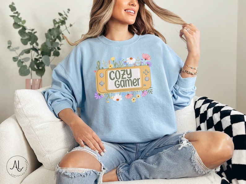 May include: A light blue sweatshirt with a graphic of a yellow video game console with the text "Cozy Gamer" surrounded by colorful flowers.