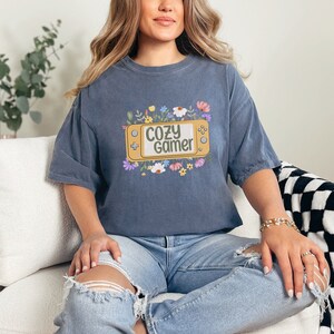 May include: A blue t-shirt featuring a graphic of a video game console with the text "Cozy Gamer" surrounded by flowers.