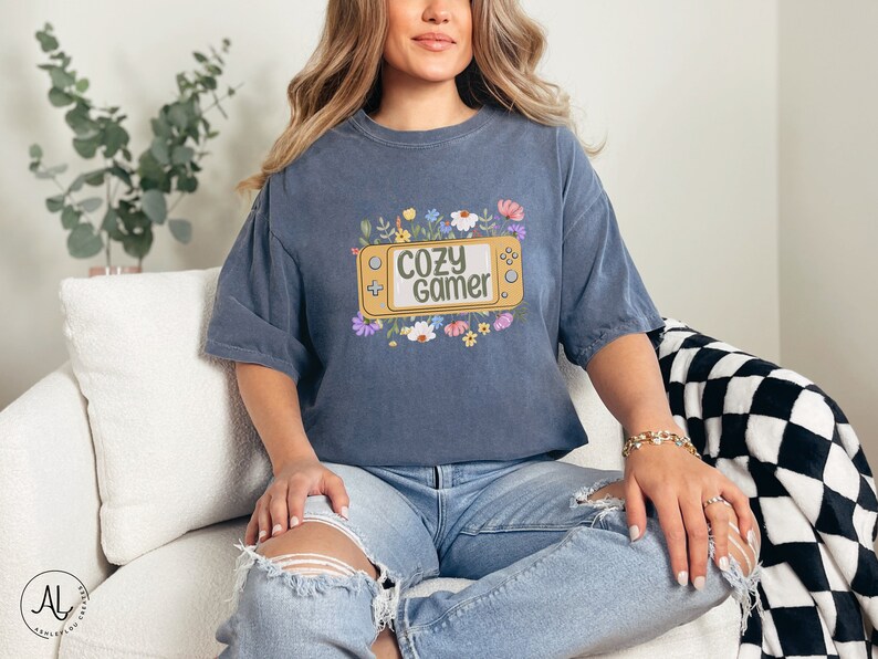May include: A blue t-shirt featuring a graphic of a video game console with the text "Cozy Gamer" surrounded by flowers.