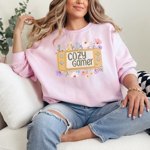 May include: A pink sweatshirt with a graphic of a video game controller surrounded by flowers. The text on the controller reads "Cozy Gamer".