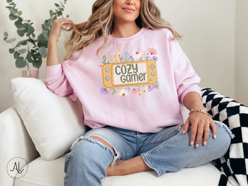 May include: A pink sweatshirt with a graphic of a video game controller surrounded by flowers. The text on the controller reads "Cozy Gamer".