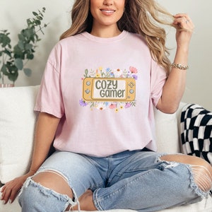 May include: A pink t-shirt featuring a graphic of a video game controller surrounded by flowers. The text "Cozy Gamer" is on the controller.