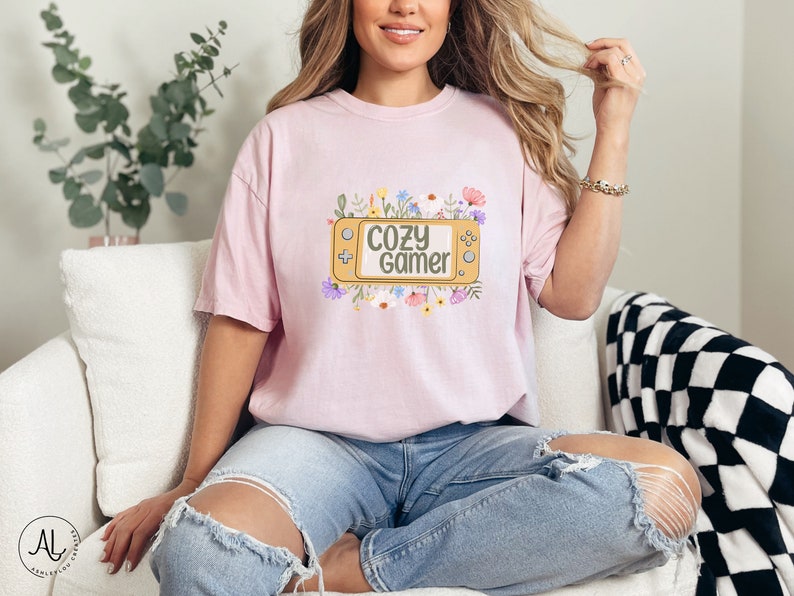 May include: A pink t-shirt featuring a graphic of a video game controller surrounded by flowers. The text "Cozy Gamer" is on the controller.