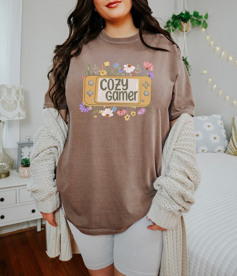 May include: A brown t-shirt featuring a graphic of a video game console with the text "Cozy Gamer" surrounded by flowers.