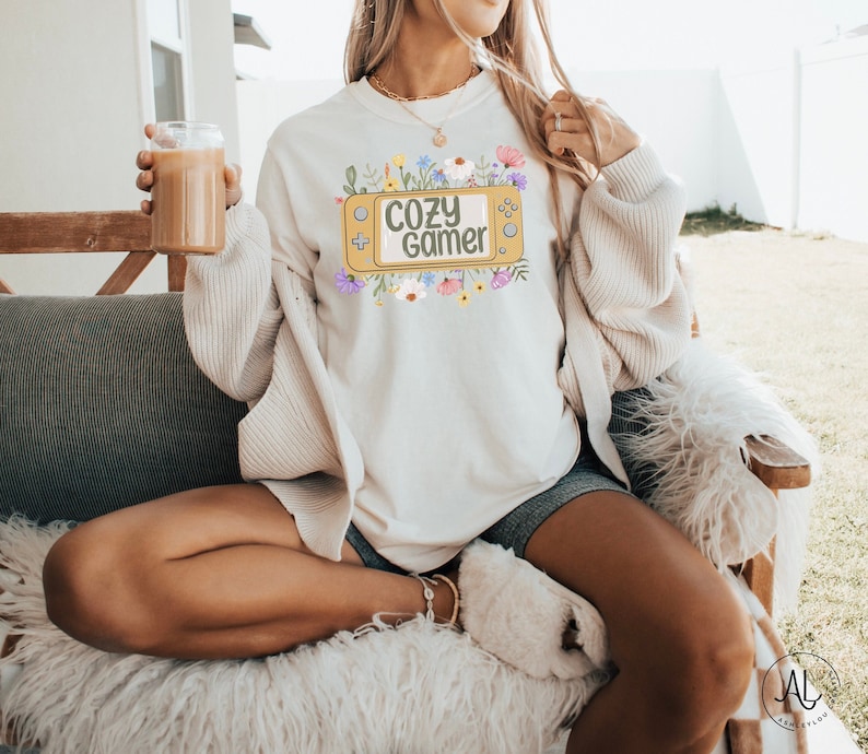 May include: A white sweatshirt featuring a graphic of a yellow video game controller with the text "Cozy Gamer" in green letters. The graphic is surrounded by colourful flowers.