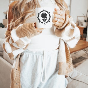 May include: A white ceramic mug with a black skull and dagger design. The mug is being held by a person wearing a white tank top and a brown and white cardigan.