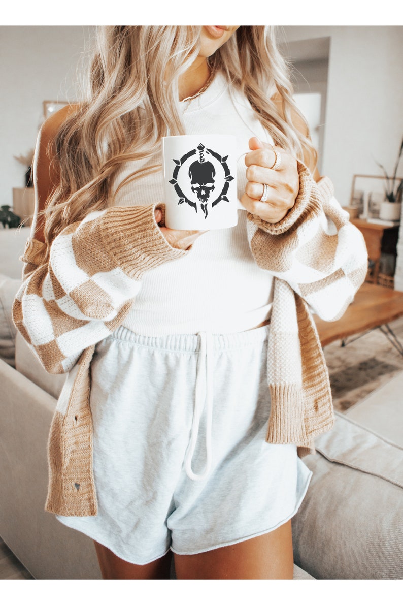 May include: A white ceramic mug with a black skull and dagger design. The mug is being held by a person wearing a white tank top and a brown and white cardigan.