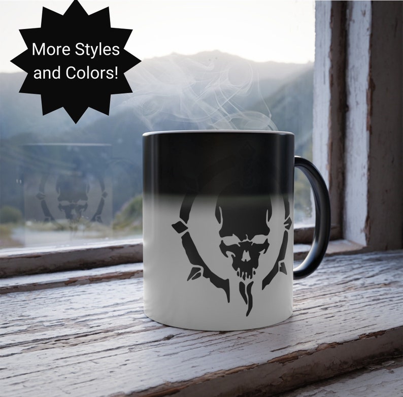 May include: Black and white color changing mug with a skull and thorns design. The mug is on a wooden surface in front of a window with a view of mountains.