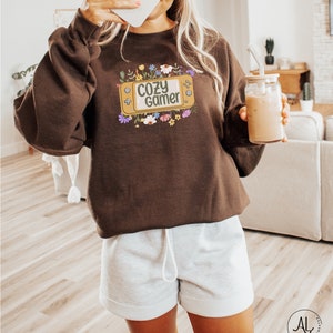 May include: A brown sweatshirt with a graphic of a video game console with the text "Cozy Gamer" surrounded by colorful flowers.