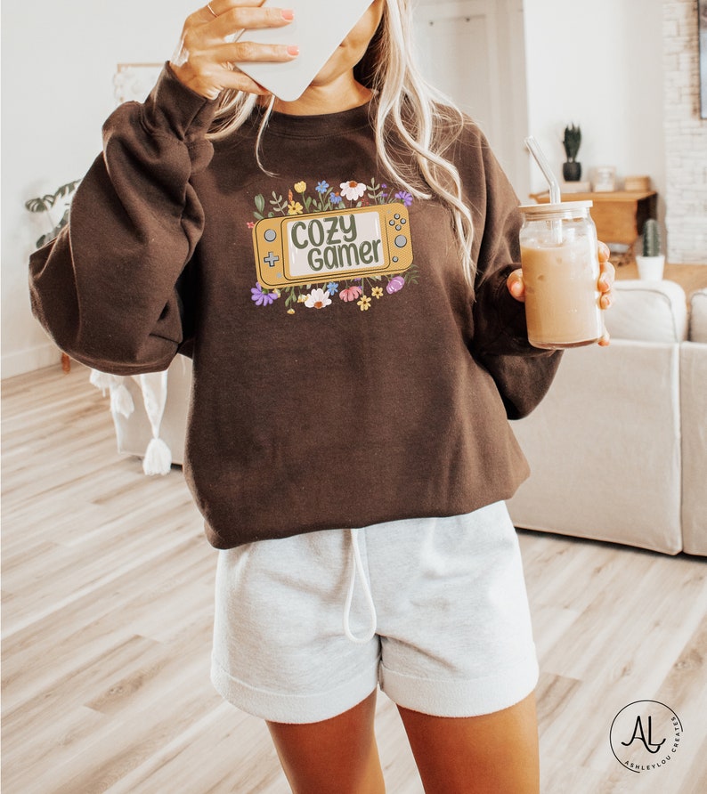 May include: A brown sweatshirt with a graphic of a video game console with the text "Cozy Gamer" surrounded by colorful flowers.