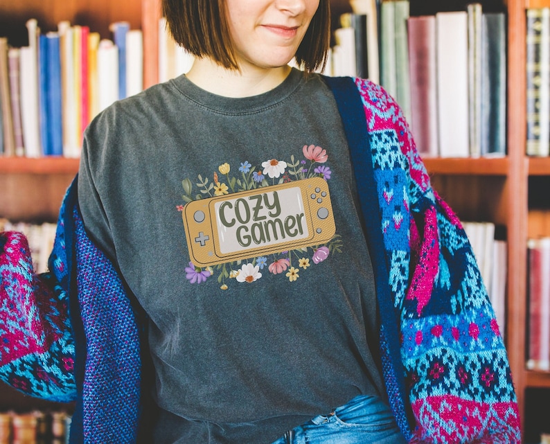 May include: A dark grey t-shirt featuring a floral design and a stylised video game controller with the text "Cozy Gamer" on it.
