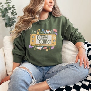 May include: A green sweatshirt with a graphic of a yellow video game console with the text "cozy gamer" surrounded by colorful flowers.