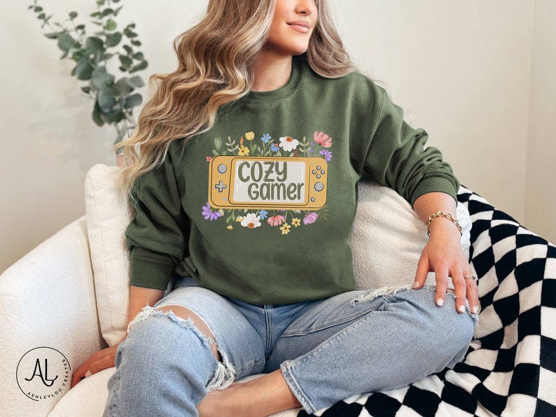 May include: A green sweatshirt with a graphic of a yellow video game console with the text "cozy gamer" surrounded by colorful flowers.