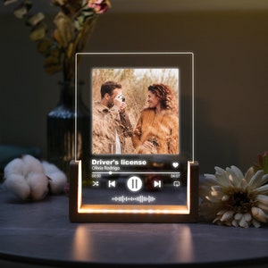 May include: A wooden photo frame with a clear acrylic insert. The frame has a built-in LED light strip that illuminates the photo and music player controls. The photo shows a couple in a field. The music player controls display the song title "Driver's license" by Olivia Rodrigo.
