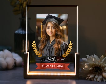 May include: A personalized graduation photo frame with a gold-colored laurel wreath and a red ribbon with the text "CLASS OF 2024". The frame has a warm white LED light around the edge and a wooden base.